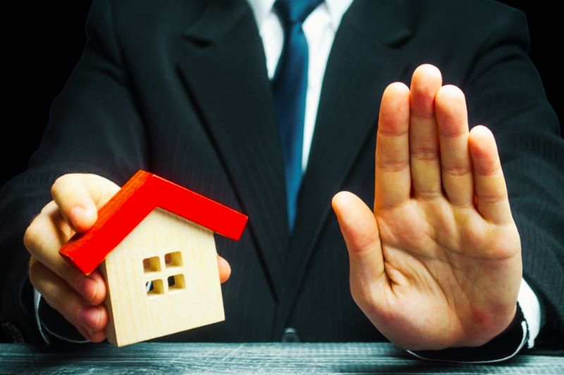 Abogado señalando con la mano en alto mientras sostiene una casa de juguete, simbolizando la protección contra fraudes inmobiliarios.